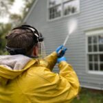 Person cleaning house siding with a pressure washer, wearing protective gear in a yellow raincoat.