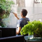 A man cleaning a glass barrier outdoors with a squeegee, surrounded by greenery.
